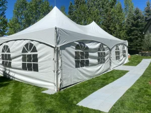 White 20x40 event tent with tall peaks and window panels, set on green lawn beside paved walkways and surrounded by leafy trees.