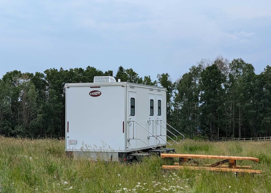 White 3-stall washroom trailer with LANG logo, side doors and metal handrails parked in a grassy field beside trees under a cloudy sky outdoors.