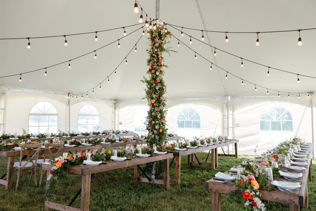Wedding Tent Packages 7 White pole tent wedding reception with rustic wooden tables, warm string lights, and a tall floral centerpiece in bright orange and pink blooms