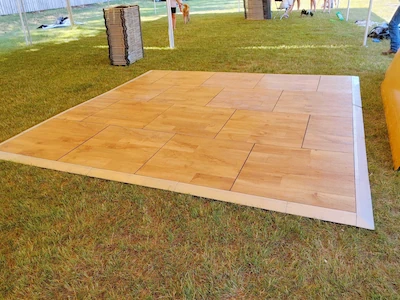 Tan wooden dance floor panels laid on green grass outdoors, bordered by clean edges, creating a smooth event walkway surface.