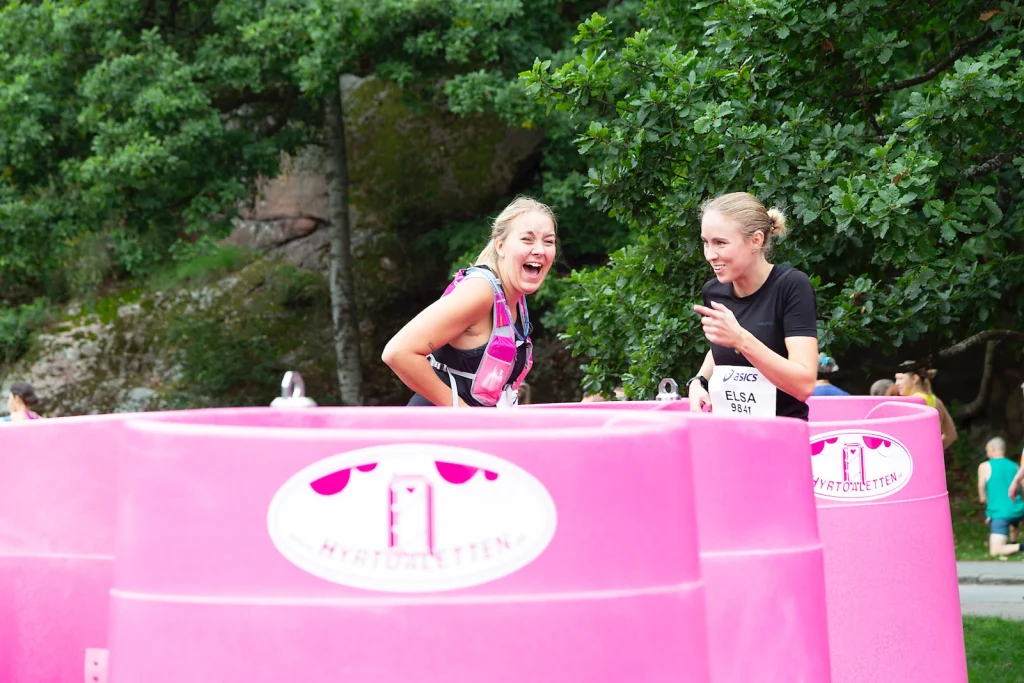 Laughing runners beside a Lapee pink portable female urinal outdoors, with green trees in the background and a race bib reading ELSA 9841.