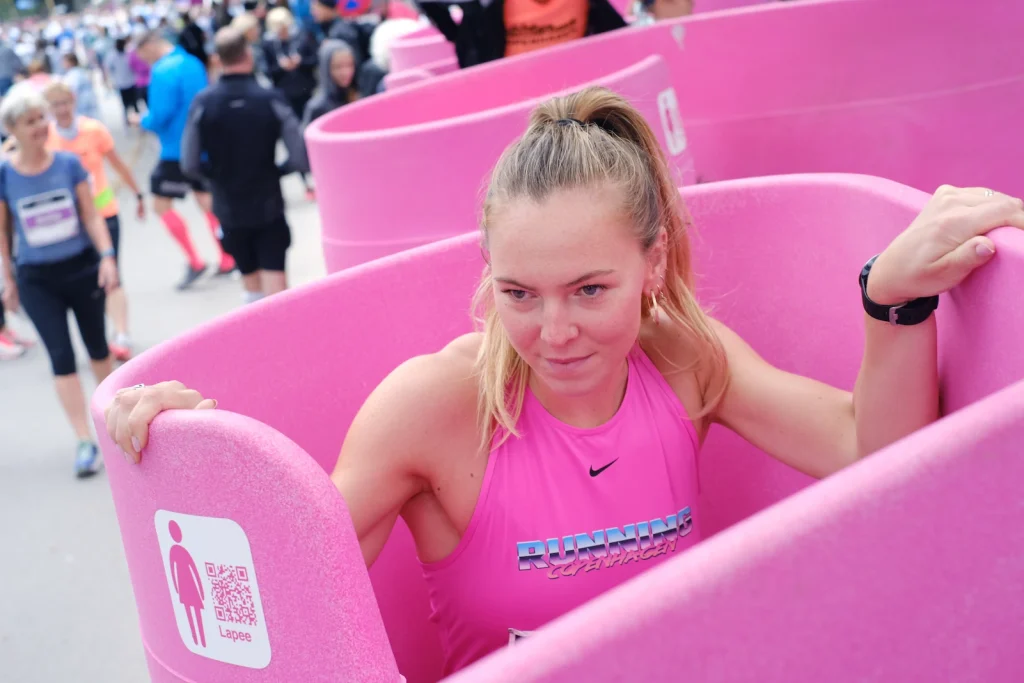 A participant in a pink sports top uses a Lapee female urinal at a busy race, with pink units and a crowd in the background.