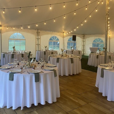 White linen-covered event tables arranged under warm string lights inside a clear-span tent with teal arched window panels and place settings.