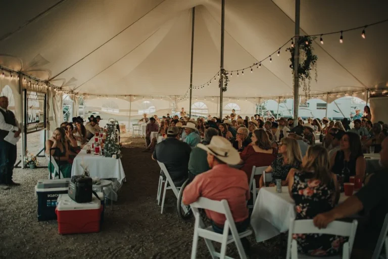 Guests seated under a white event tent during a wedding reception, with string lights above and reception tables set for celebration.