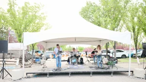 Outdoor event stage under a white canopy with musicians performing, guitars and keyboards at the ready, and green trees surrounding the venue area.