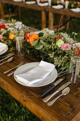 Wooden event table set with folded white napkins, silver cutlery, and bright orange and pink florals in glass vases, outdoors.
