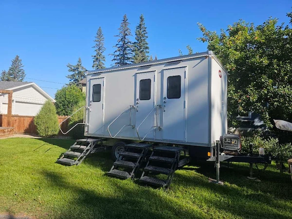 Wedding Tent Packages 15 White and blue washroom trailer with exterior access steps on a green lawn, set in an outdoor farm yard under a clear blue sky.