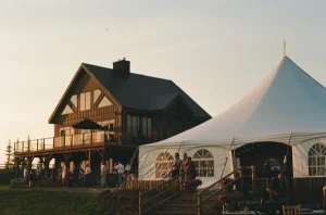 Dusk lighting over blue-and-white rental tents and a silhouetted lodge house, with guests gathered in the open event area.
