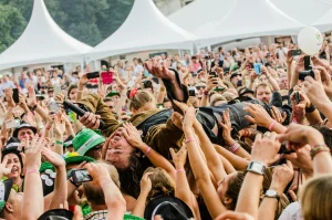 A lively outdoor crowd raises phones and hands in front of white event tents, capturing the moment onstage with excitement.