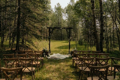 Wooden ceremony chairs line a forest aisle under an arch, with soft pathway lighting and lush green trees surrounding the setup
