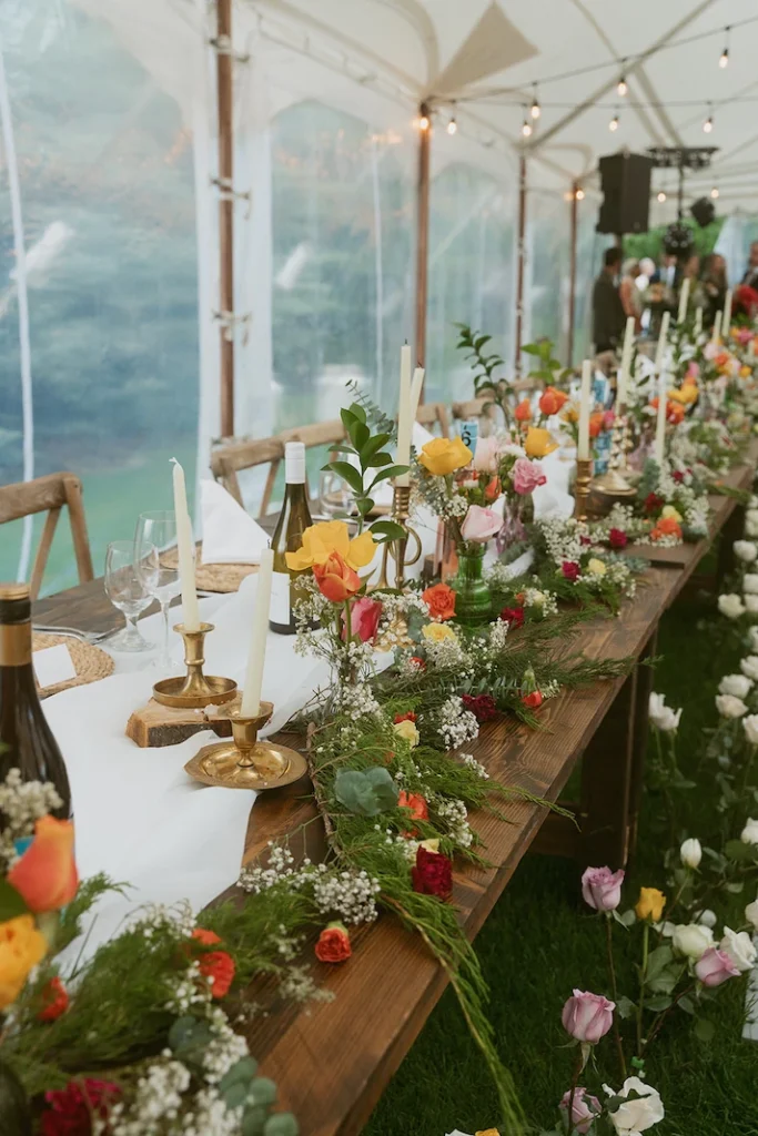 Wedding Tent Packages 9 Rustic wooden event tables in an elegant sailcloth tent, lined with brass candle holders and vibrant mixed flower arrangements.