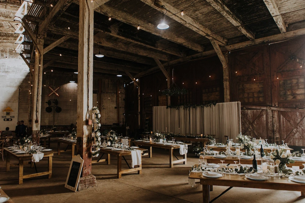 Rustic wedding setup inside an old railway shed with wooden beams, warm string lights, long tables, floral centrepieces, and white draping.