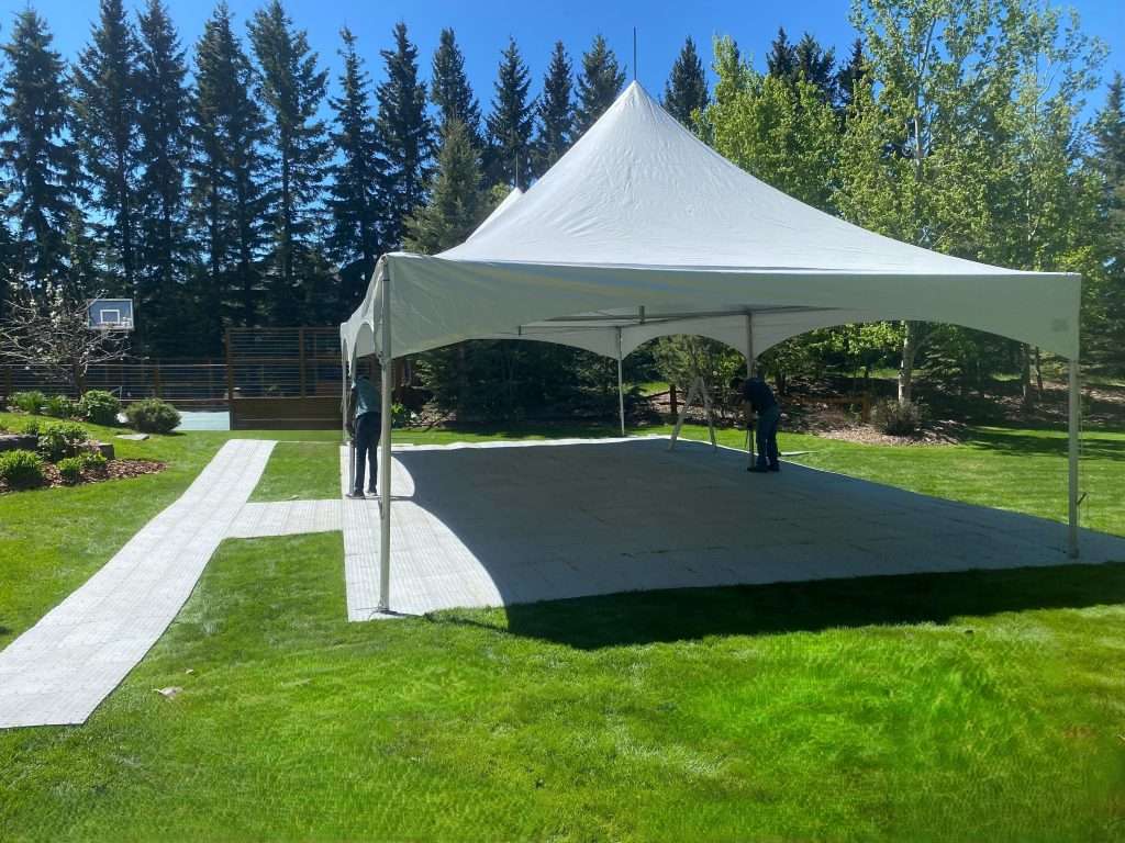 Backyard Tent Rental In Calgary Ab | Vibe Event Rentals 7 White 20x40 backyard tent stands on a manicured Calgary lawn as two installers work inside, with a walkway, trees, and clear blue sky around it.
