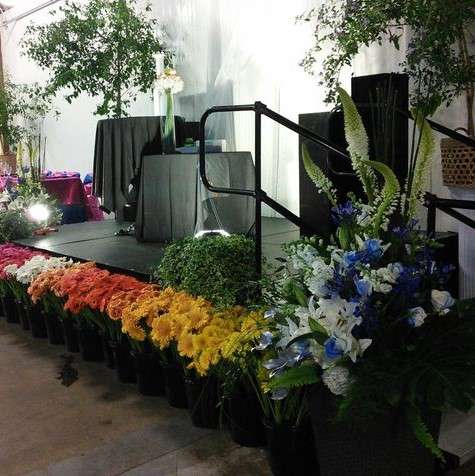 A black modular stage with railings is set among floral arrangements and greenery at an indoor venue, ready for a Calgary event on uneven flooring.