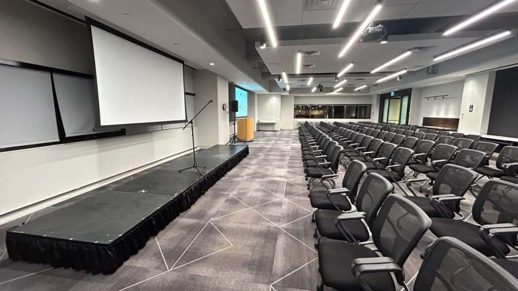 A low black stage with skirting stands at the front of a modern conference room, beside a projection screen and rows of black event chairs.